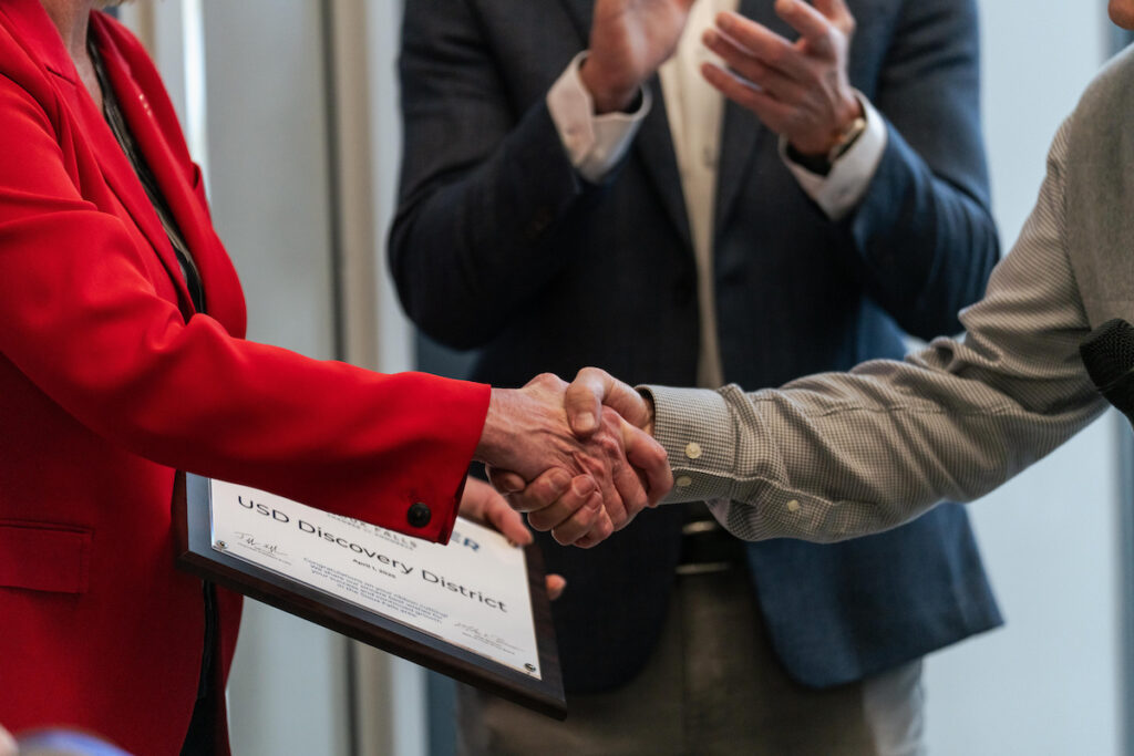 Close-up of USD President Sheila Gestring shaking hands with an attendee while holding a framed certificate at the USD Discovery District Grand Opening on April 1, 2025.