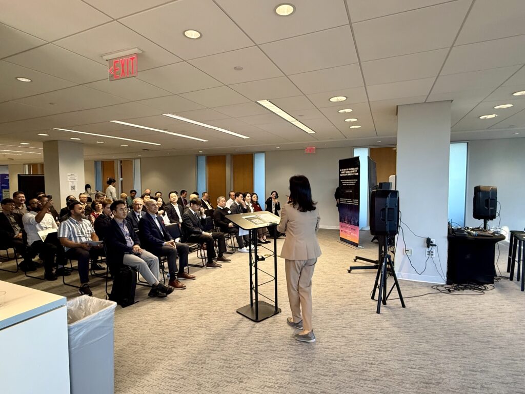 Julie Koo, President and CEO of Luceque Global, stands at the front of a conference room speaking to a full audience during the K-Startup Showcase in Tysons, Virginia.