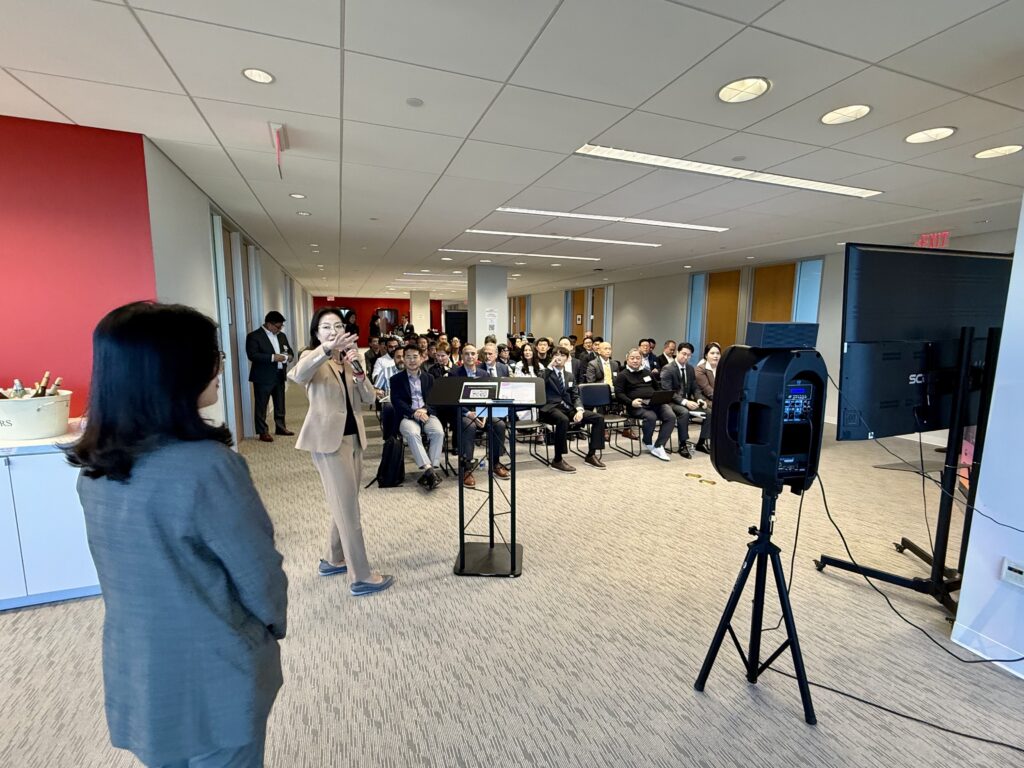 Julie Koo, President and CEO of Luceque Global, stands at the front of a conference room speaking to a full audience during the K-Startup Showcase in Tysons, Virginia.