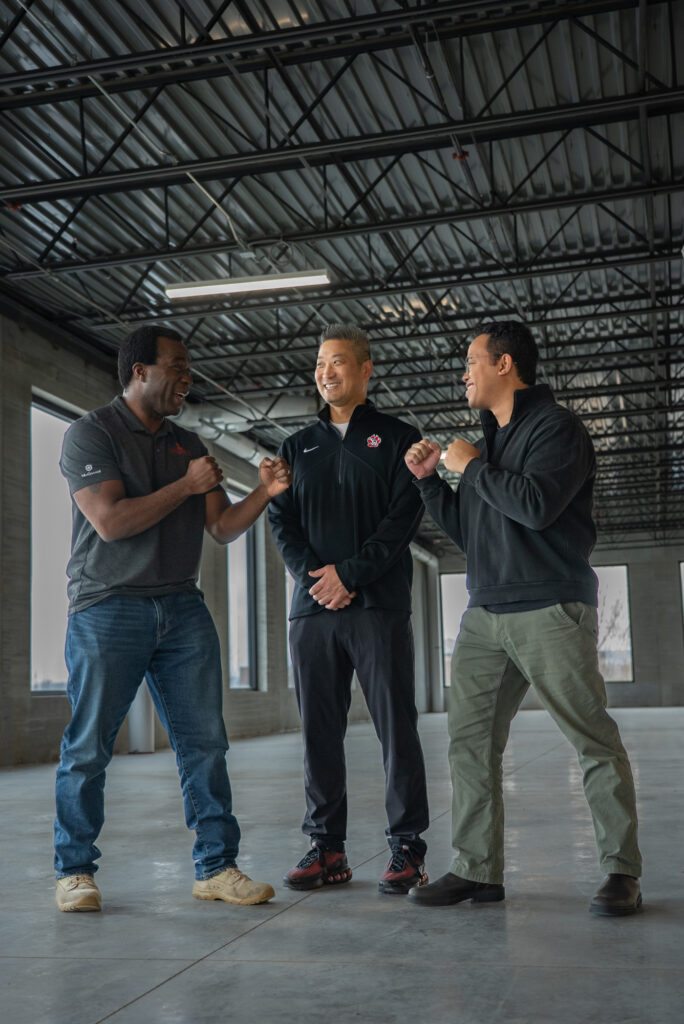Marc and two colleagues smiling and playfully posing in a mock boxing stance inside a large commercial building.